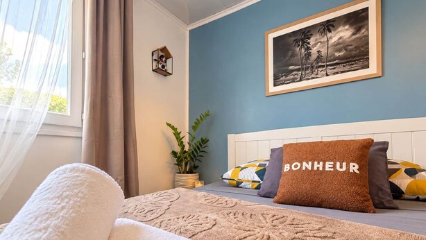 A bedroom with a bed, a pillow with 'BONHEUR' text, a framed picture on the wall, and a window with curtains. - Petit Eden du Bras Piton (Chalet) (La Plaine-des-Palmistes)