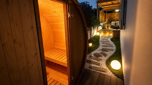 Una estructura de sauna de madera con puerta abierta, un camino de piedra y una zona de estar con una mesa y sillas.