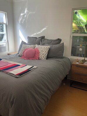 Ein Schlafzimmer mit Bett, Kissen, Nachttisch, Lampe und einem Fenster mit Blick auf Grünflächen.