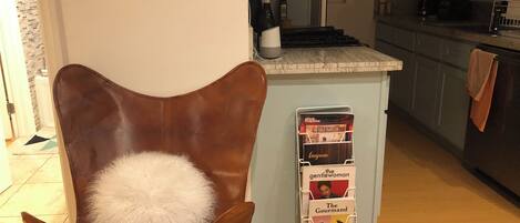 A brown leather chair with a white fluffy pillow, a magazine rack with 'The Gentlewoman' and 'The Gourmand' magazines, a kitchen area with a countertop, and a tiled floor.