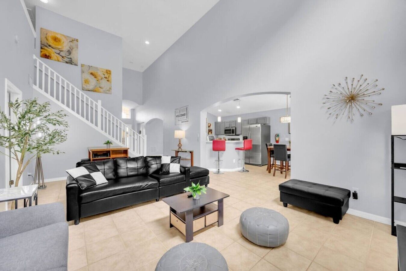 A spacious living area with a black leather sofa, a coffee table, and a grey ottoman. A staircase leads to an upper level, and a kitchen is visible in the background.