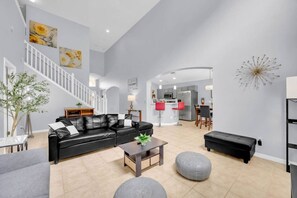 Ein geräumiger Wohnbereich mit einem schwarzen Ledersofa, einem Couchtisch und einem grauen Hocker. Eine Treppe führt auf eine obere Ebene, und im Hintergrund ist eine Küche zu sehen.