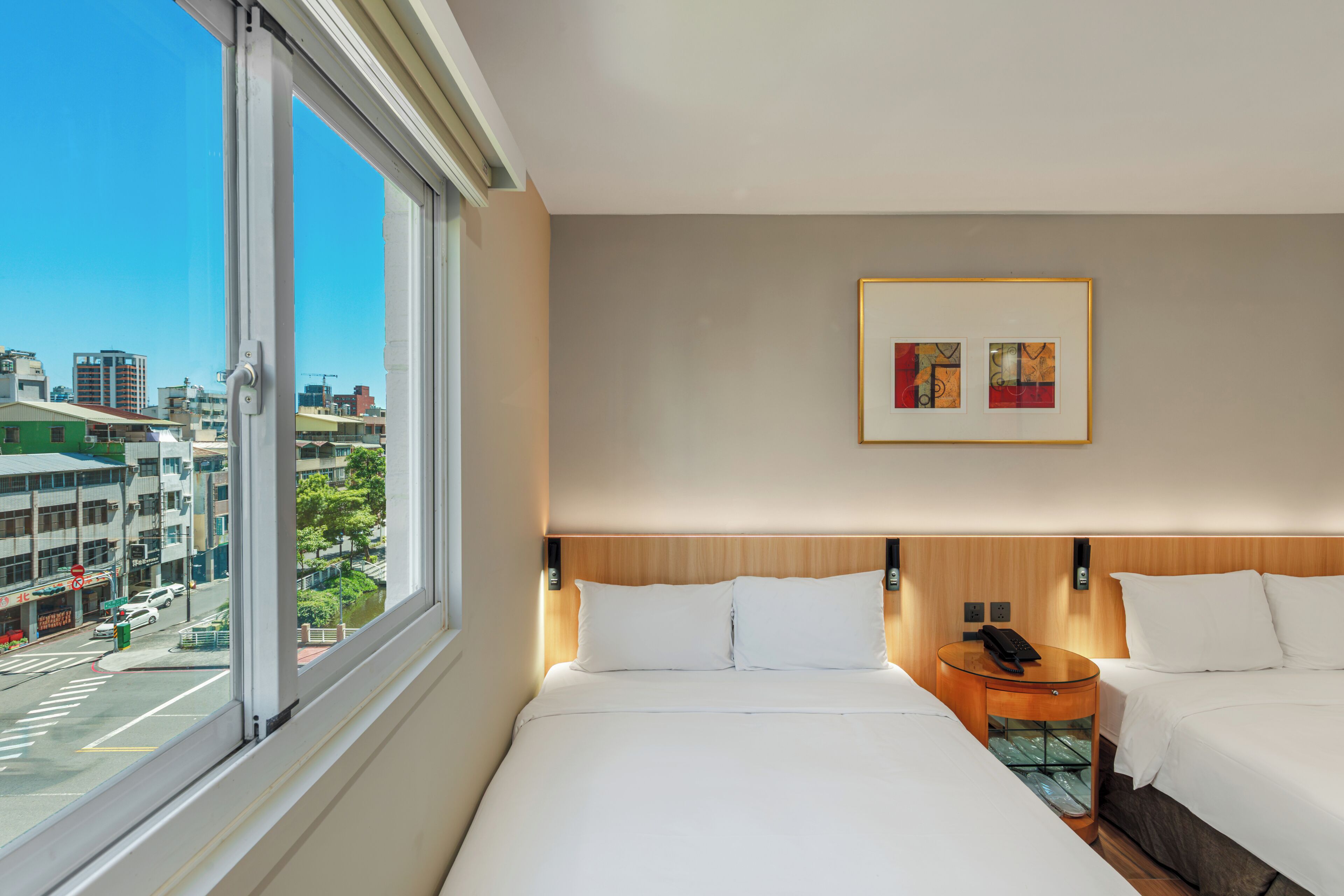 A hotel room with a large window, a bed, a bedside table with a phone, and a framed picture on the wall.