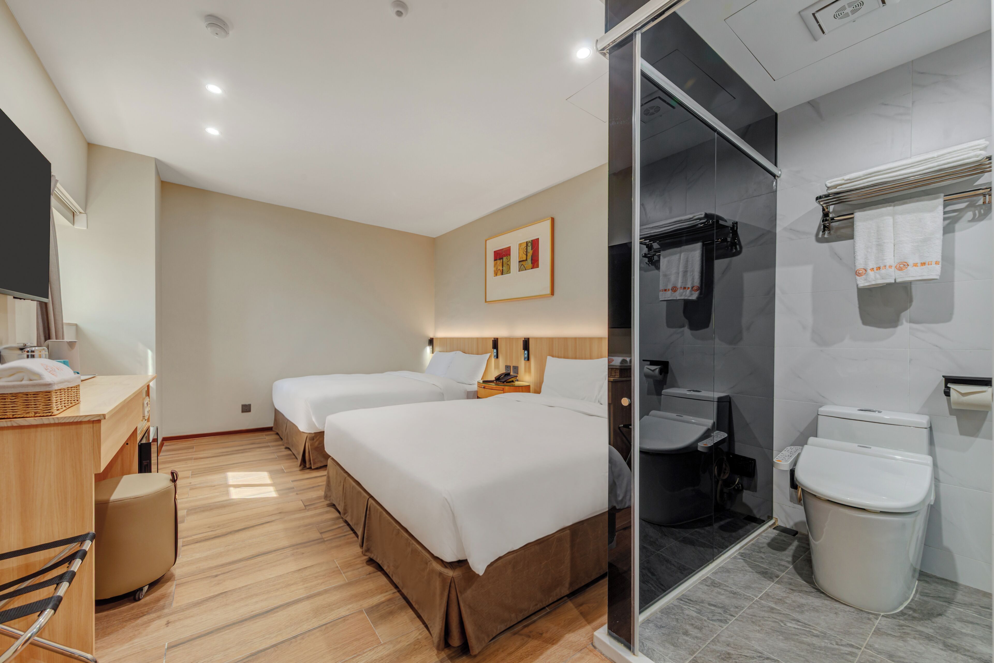 A hotel room with a wooden desk, two beds, a bathroom with a toilet and shower, and a framed picture on the wall.