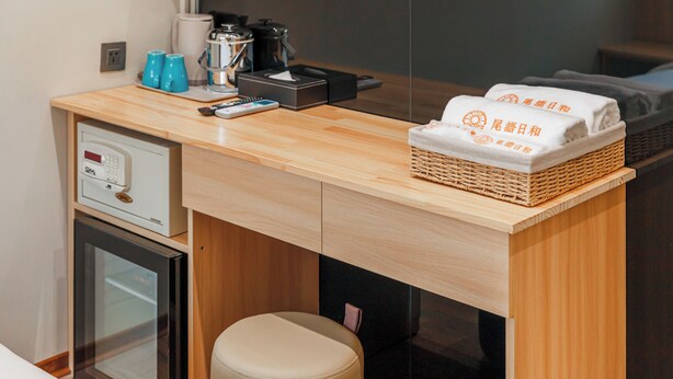A hotel room with a wooden desk, a minibar, a small refrigerator, a cushioned stool, and a woven basket with towels. - WHIYORI INN (Kaohsiung)