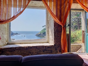 Ein Zimmer mit großem Fenster, das auf eine felsige Küstenlinie und den Blick auf das Meer blickt, sowie eine Steinsäule.