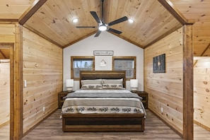Un dormitori de cabana de fusta amb un llit, dues taules de nit, un ventilador de sostre i una finestra.