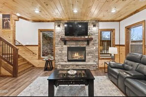 A living room with a stone fireplace, a flat-screen TV, a sofa, and a foosball table. - New Cabin Near Gatlinburg & Dollywood (Sevierville)