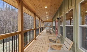 A covered wooden deck with benches, railings, and windows overlooking a forested area. - New Cabin Near Gatlinburg & Dollywood (Sevierville)