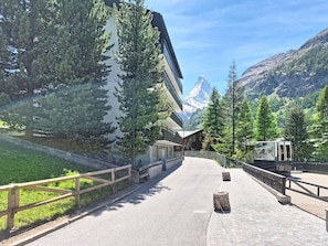 Ein gepflasterter Weg, der von Holzwailen gesäumt wird, ein Gebäude mit moderner Architektur und ein Berg im Hintergrund.