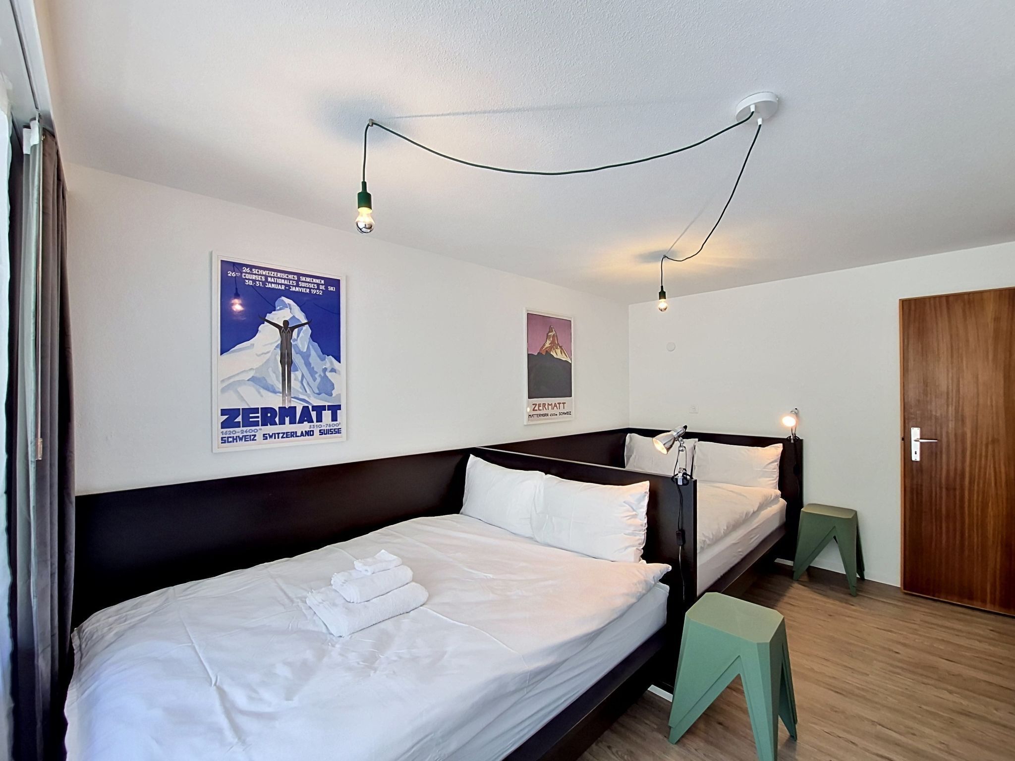 A hotel room with two beds, a wooden floor, a green stool, and a poster on the wall.
