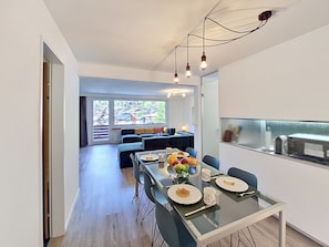 Ein moderner Essbereich mit einem Glastisch, Stühlen und Blick auf einen Balkon mit einer Couch.