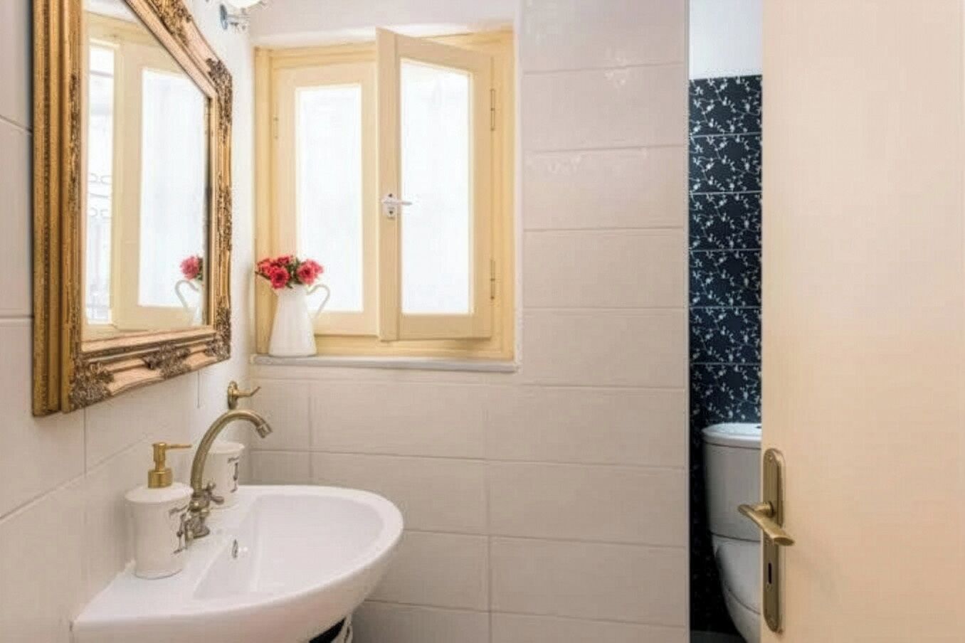 Un baño con un lavamanos blanco, un espejo con marco ornamentado, una ventana con vistas y una pared de azulejos negros.