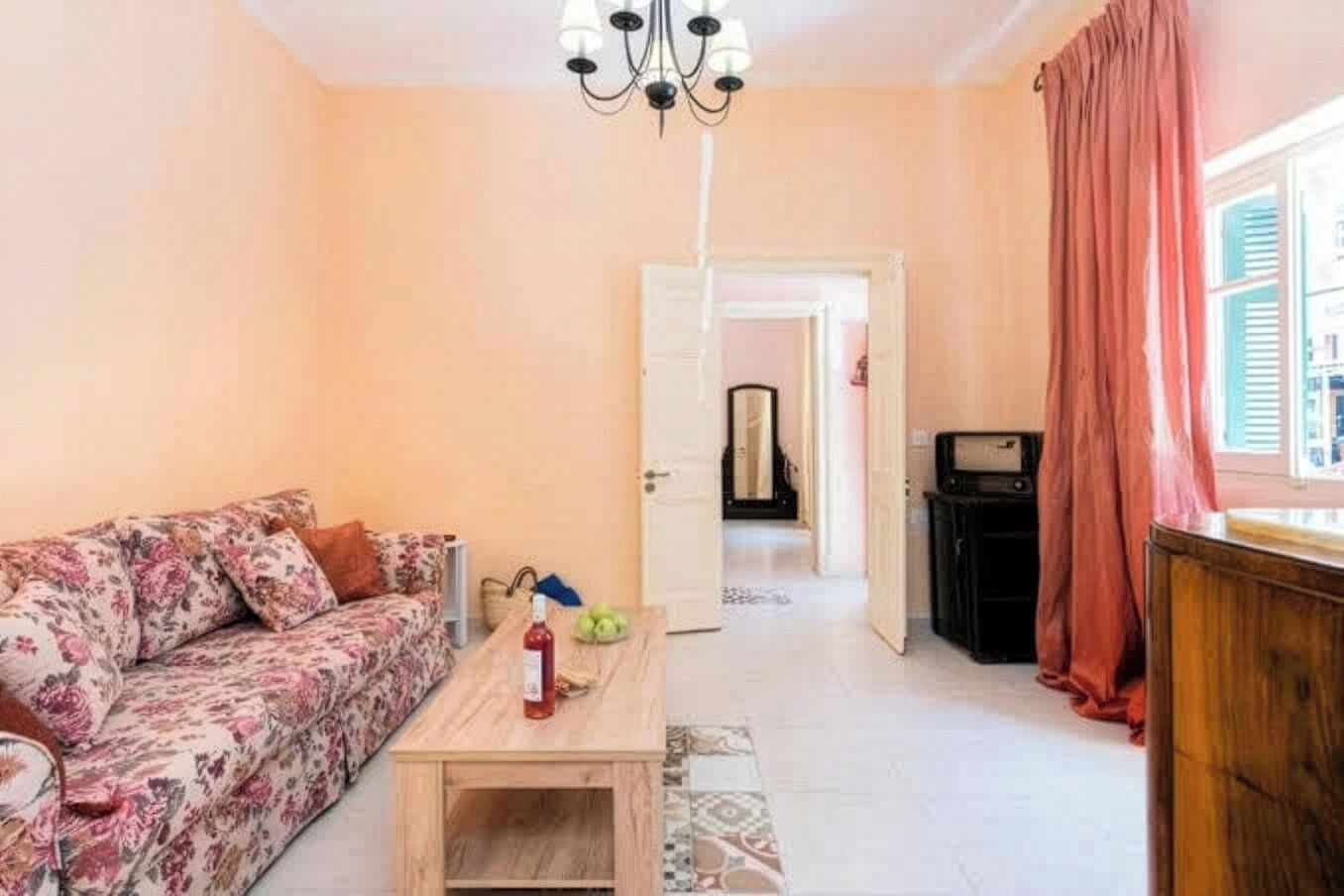 Ein Wohnzimmer mit einem blumigen Sofa, einem Holztisch, einer Chandelieber und Blick durch eine offene Tür.