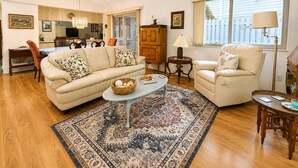 A living room with a sofa, armchair, coffee table, and a rug. There is a dining area with chairs and a table, and a kitchen in the background. - Seawoods Beach Retreat - Walk to Beach + Pools (New Smyrna Beach)
