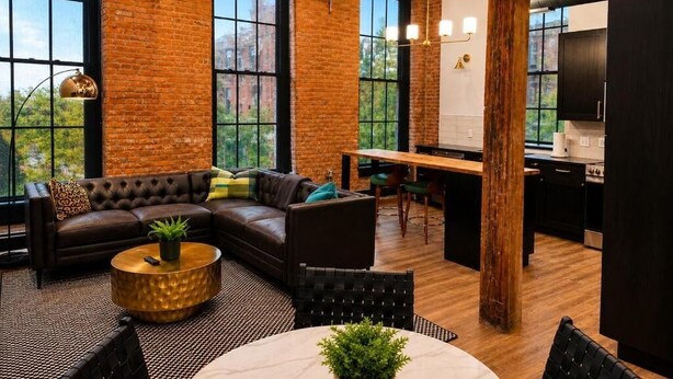 An open-plan living space with a sofa, coffee table, and dining area, featuring exposed brick walls and large windows. - Brick & Bed - Arch, Riverfront, Convention Center, T-303 (St. Louis)