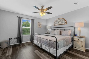 Ein Schlafzimmer mit Bett, Nachttisch, Deckenventilator, einem Fenster mit Vorhängen und einem Wandbild.
