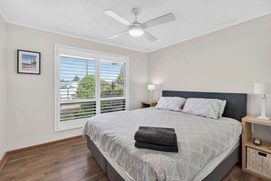 Ein Schlafzimmer mit Bett, Deckenventilator, Fenster mit Jalousien, Nachttisch mit Lampe und einem gerahmten Bild an der Wand.