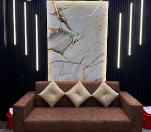 A brown sofa with three cushions and a wall with a marble-like pattern and vertical light fixtures. - Belpatra Paying Guest House (Varanasi)