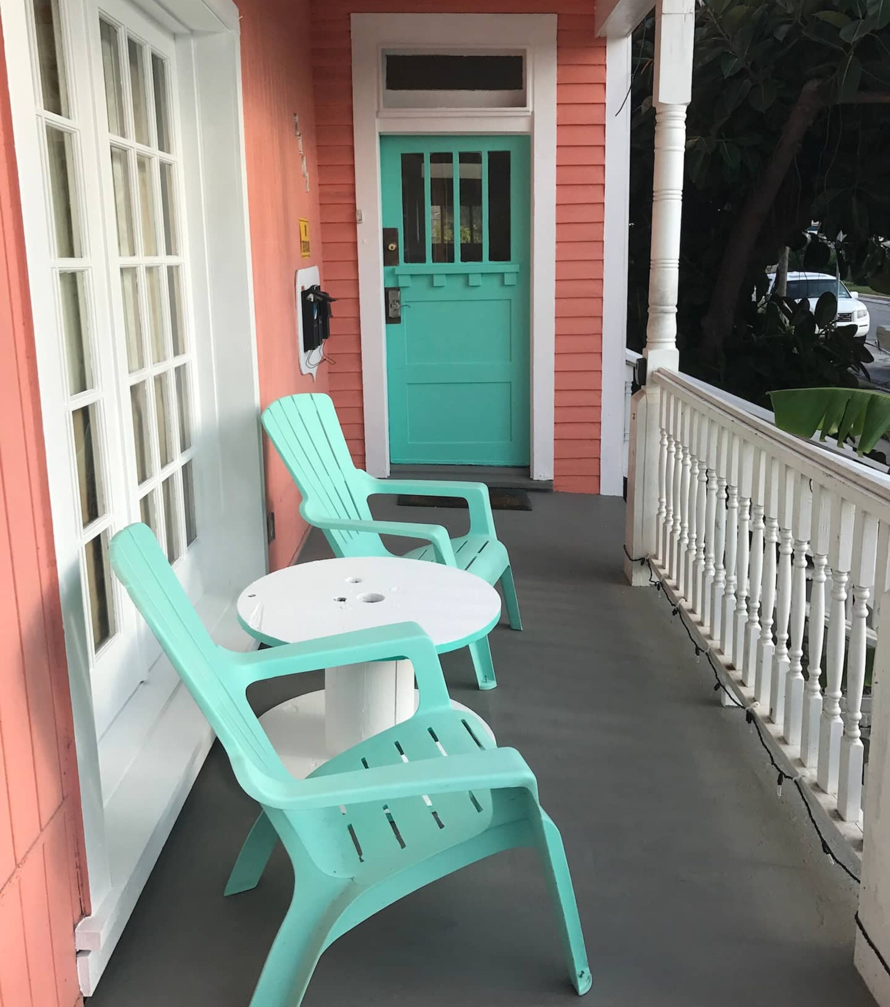 A porch with two turquoise chairs and a small table, a turquoise door, and a white railing.