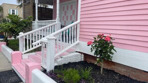 Una casa rosa con un porche blanco, escalones y un pequeño jardín con plantas.
