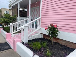 Uma casa rosa com varanda branca, degraus e um pequeno jardim com plantas.