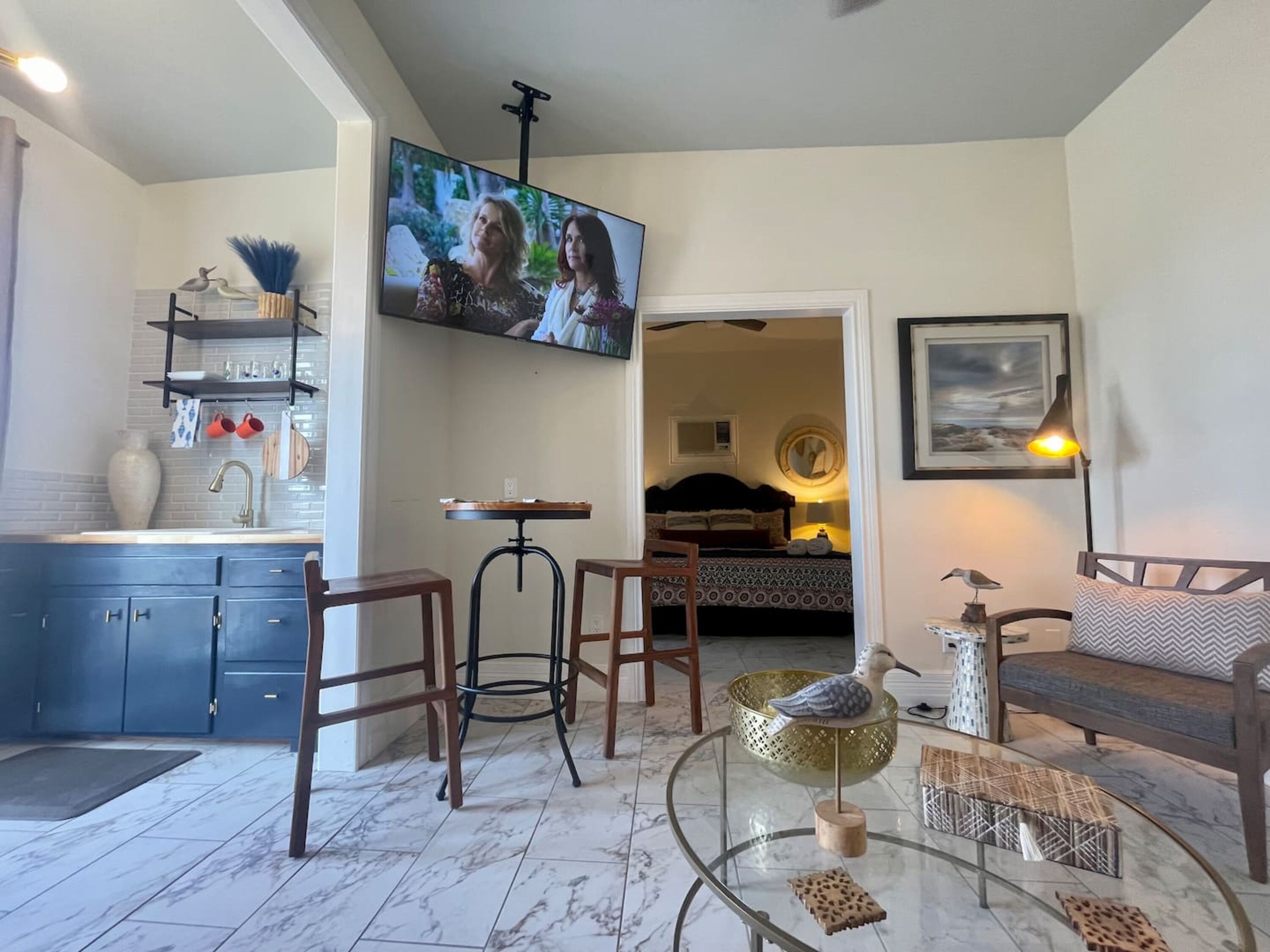 Un salon moderne avec un téléviseur, un coin repas avec des tabourets, une cuisine avec des meubles bleus et une chambre à coucher visible à travers une porte ouverte.