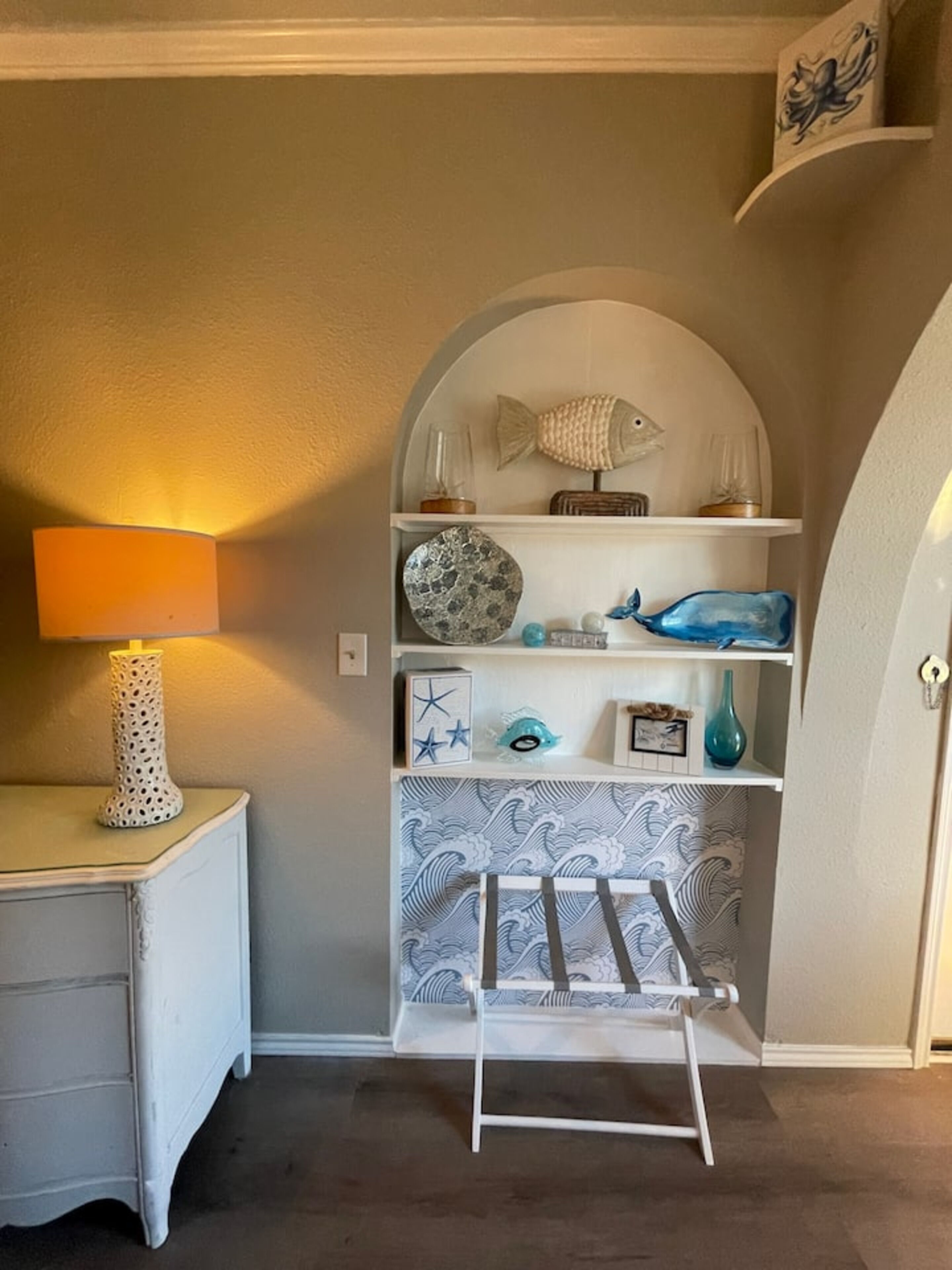 A room with a white dresser, a lamp, a shelf with decorative items, and a white drying rack.