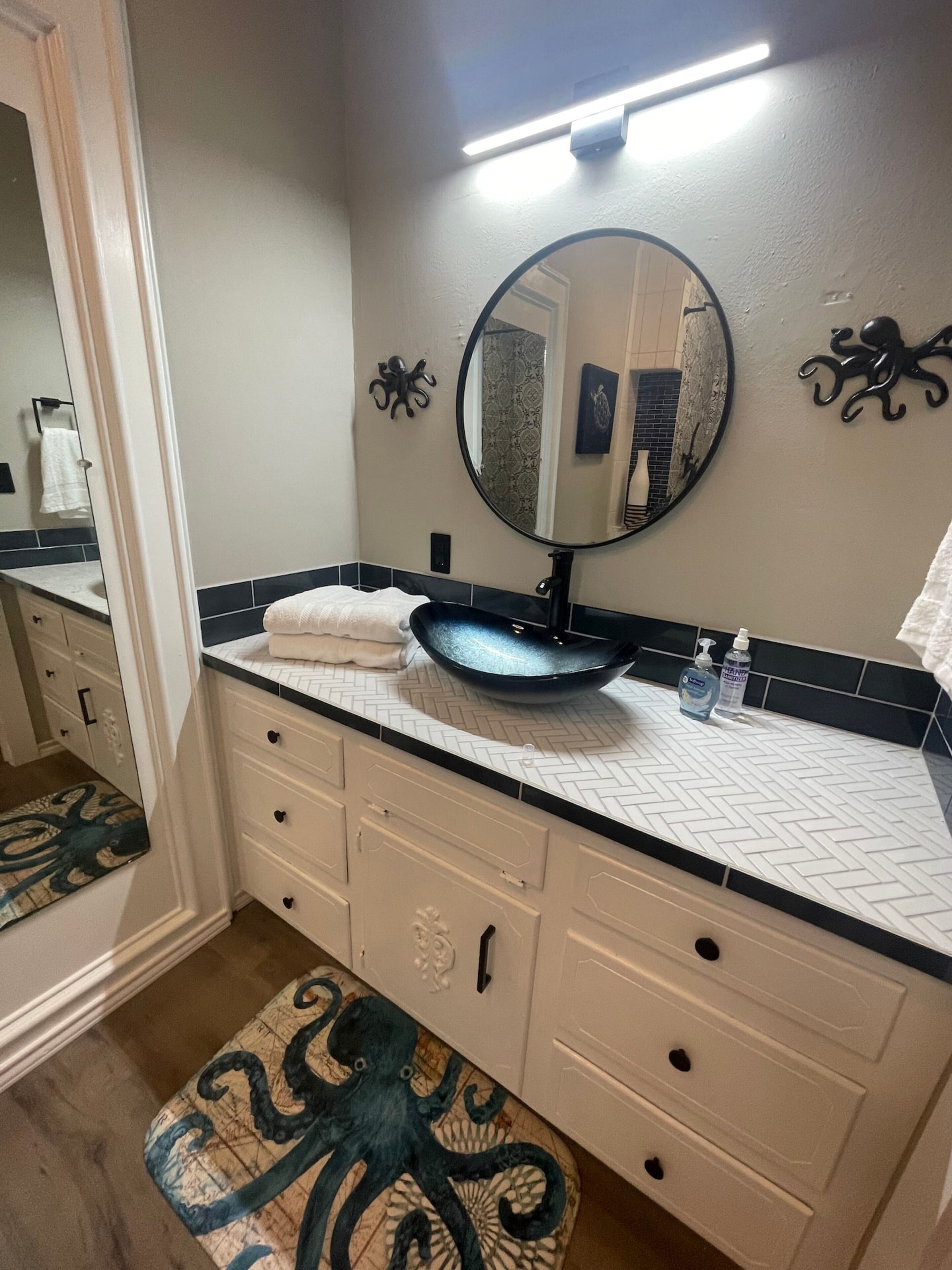 A bathroom with a round mirror, a sink with a blue basin, a towel on the counter, and a decorative octopus rug.