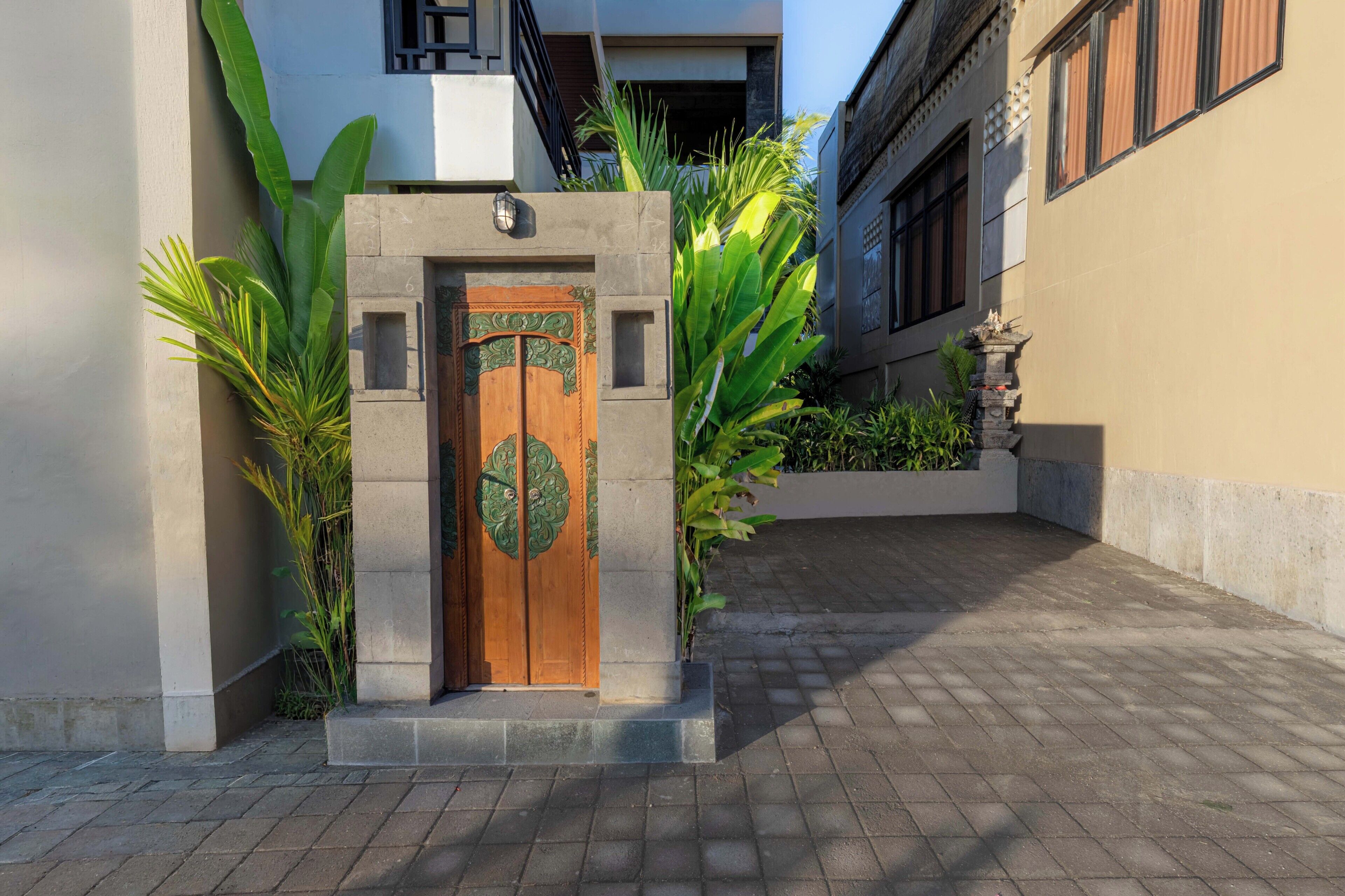 A gated entrance with a wooden door, flanked by large green plants, leading to a paved walkway.