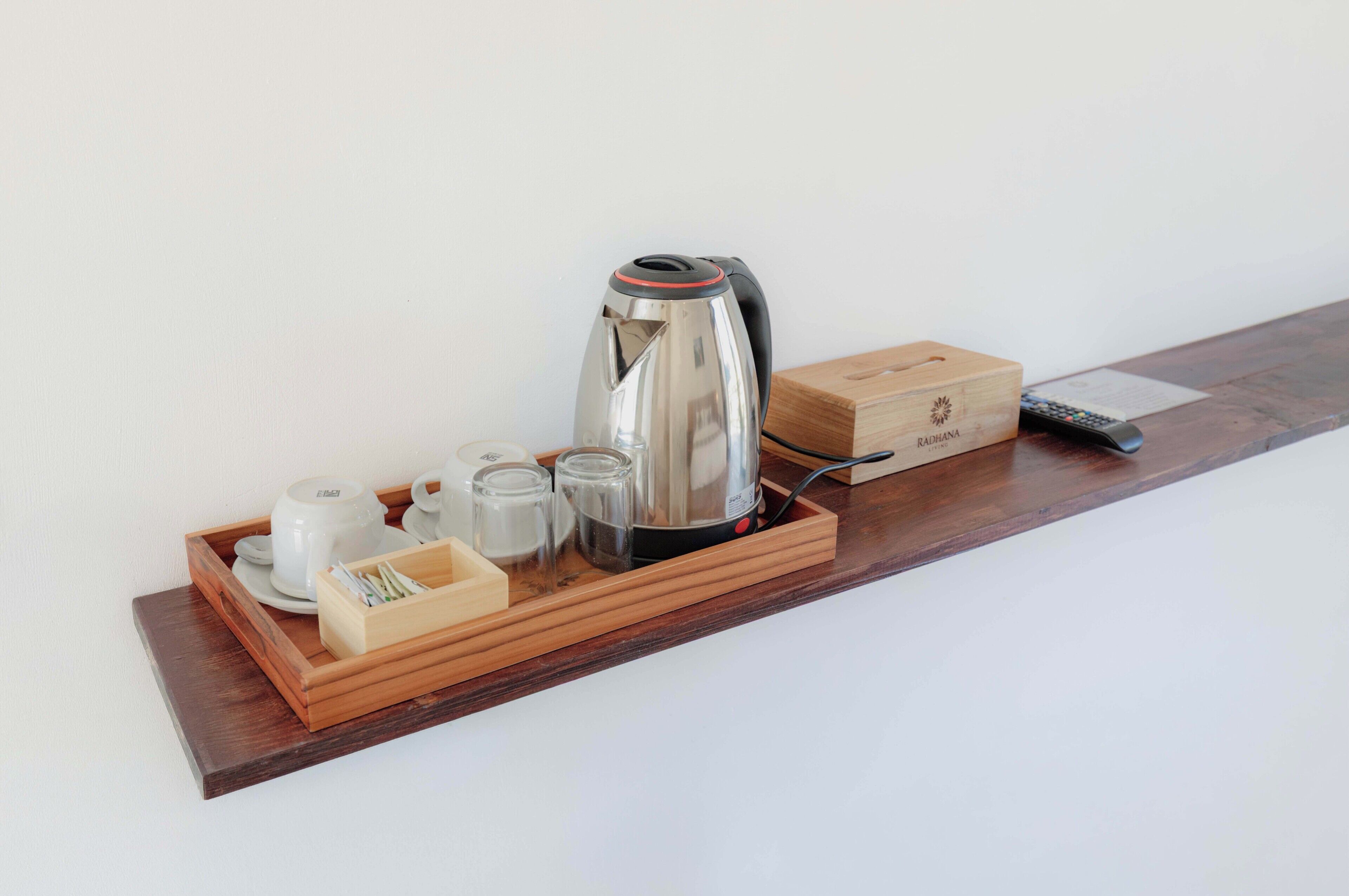 A wooden tray with a kettle, teapot, cups, and a box labeled 'Radhana' on a shelf.