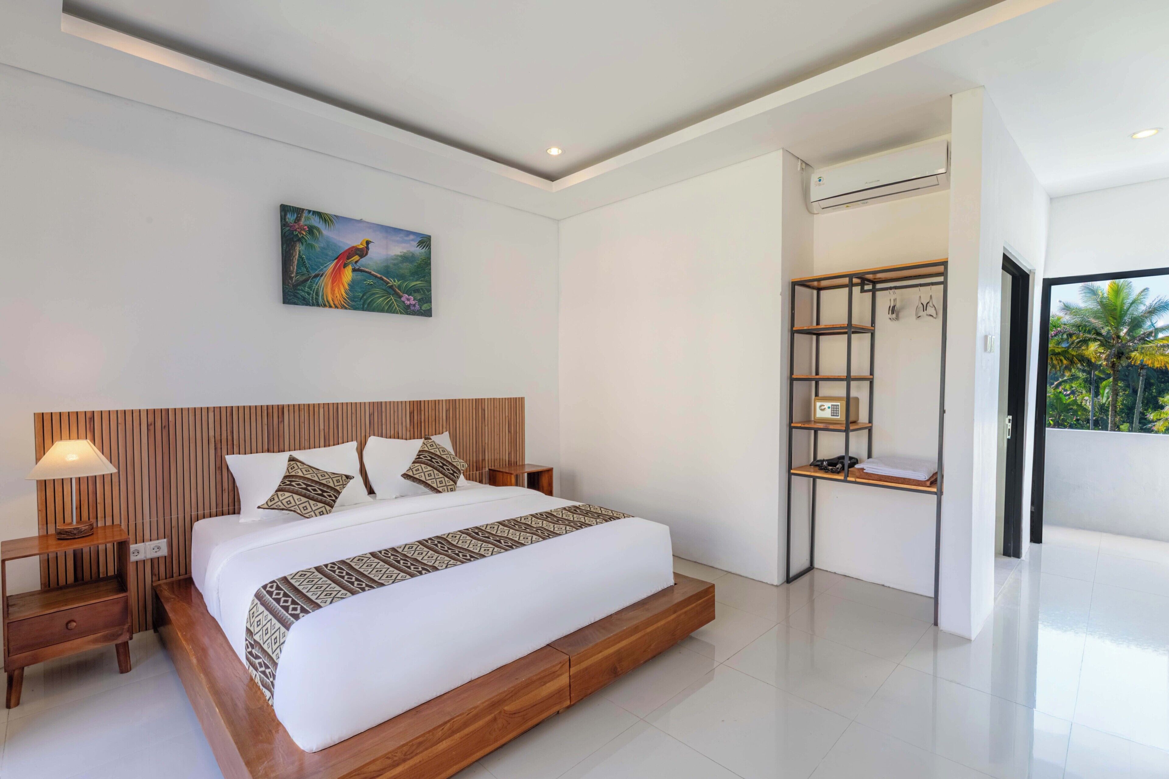 A modern bedroom with a wooden headboard, a large bed with patterned bedding, a nightstand with a lamp, and a painting of a tropical scene.