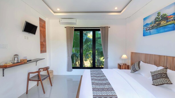 A hotel room with a bed, a desk with a chair, a TV, an air conditioning unit, and a painting on the wall. - Radhana Living (Tampaksiring)