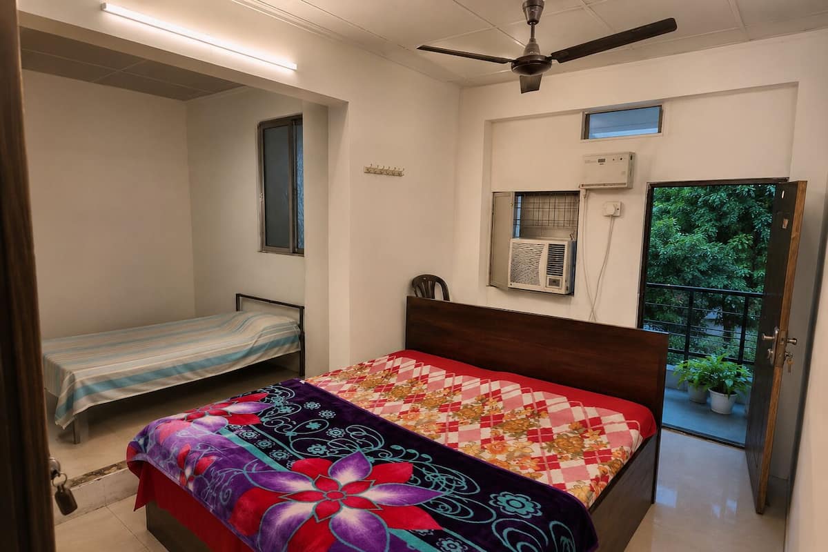 Un dormitorio con una cama individual, ventilador de techo, unidad de aire acondicionado y ventana con vista a follaje.