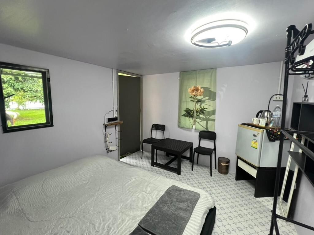 A room with a bed, a desk, two chairs, a small table, a radiator, and a window with a view of greenery.