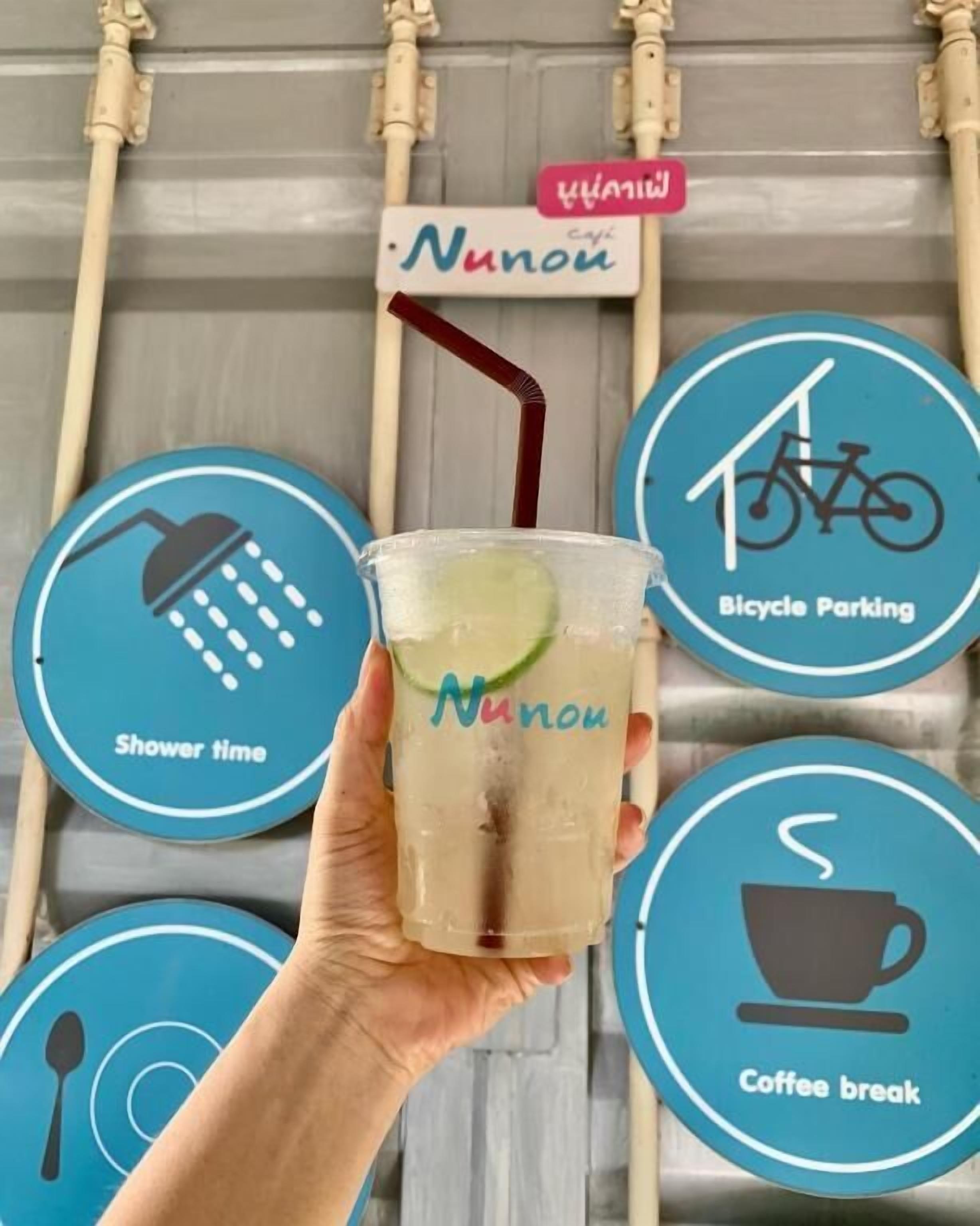 Una mano sosteniendo un vaso con pajita, un letrero que dice 'Nunou' y señales circulares que indican horario de ducha, estacionamiento para bicicletas y pausa para café.