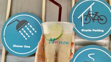Una mano sosteniendo un vaso con pajita, un letrero que dice 'Nunou' y señales circulares que indican horario de ducha, estacionamiento para bicicletas y pausa para café.