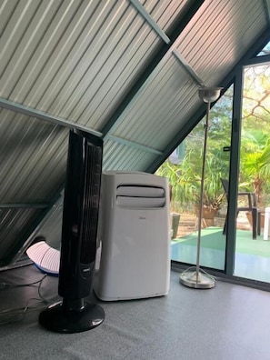 Quarto com teto inclinado, aquecedor portátil, ar condicionado branco e vista para o verde lá fora.