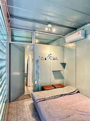 Um quarto pequeno e reservado, com uma cama, uma mesinha, ar condicionado fixado na parede e uma porta que dá para outro cômodo.