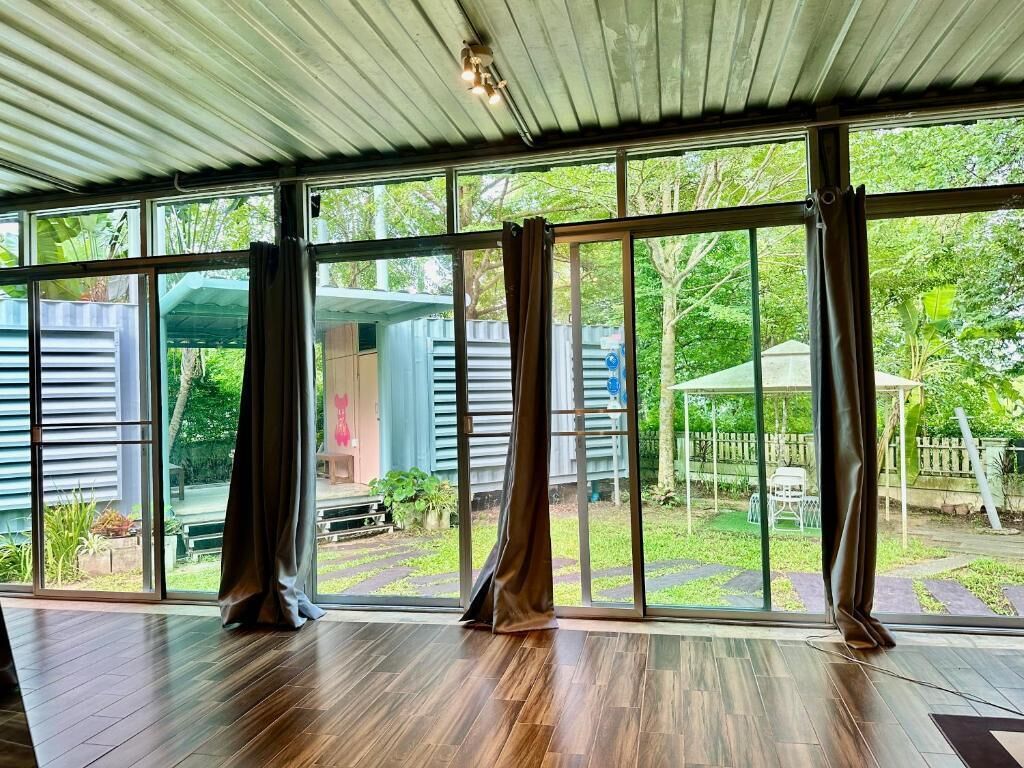 A room with large windows, a wooden floor, and a view of a garden and buildings outside.