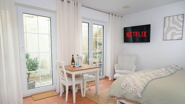 Un dormitorio con cama, televisor montado en la pared, mesa de comedor con sillas y ventana con cortinas.