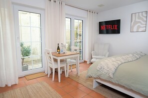 A bedroom with a bed, a TV mounted on the wall, a dining table with chairs, and a window with curtains.