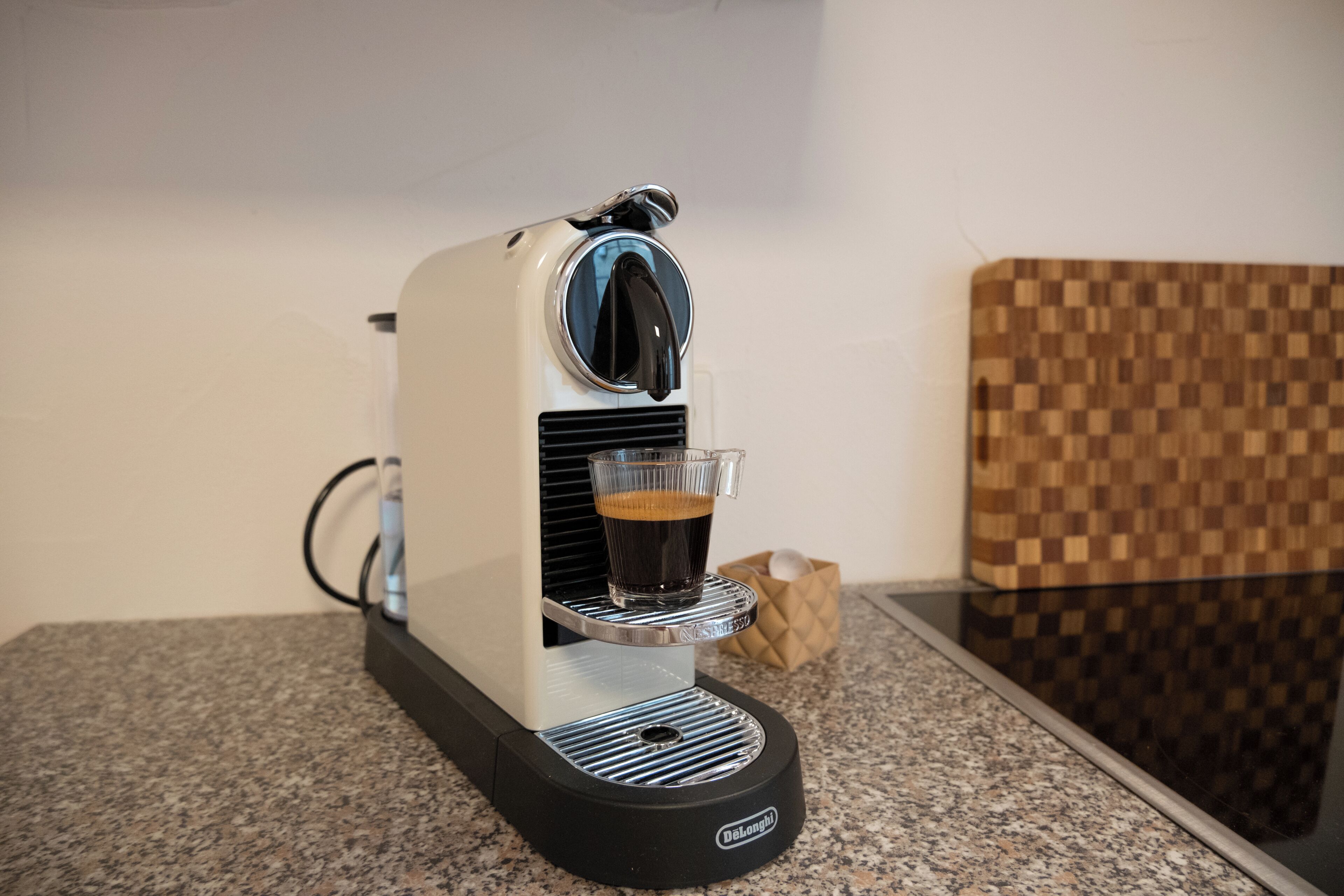 A coffee machine with a glass cup on its drip tray, a power cord, and a checkered wall in the background.