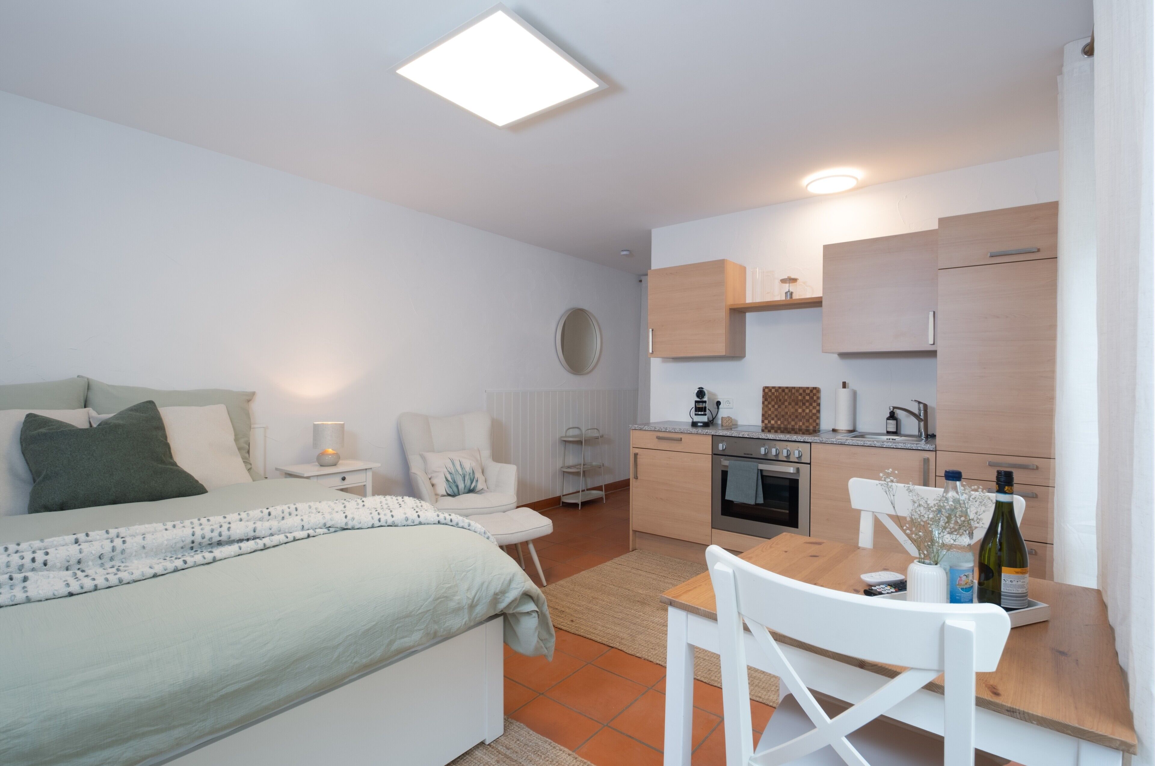 A compact room with a bed, a small dining table, a kitchenette, and a white chair.