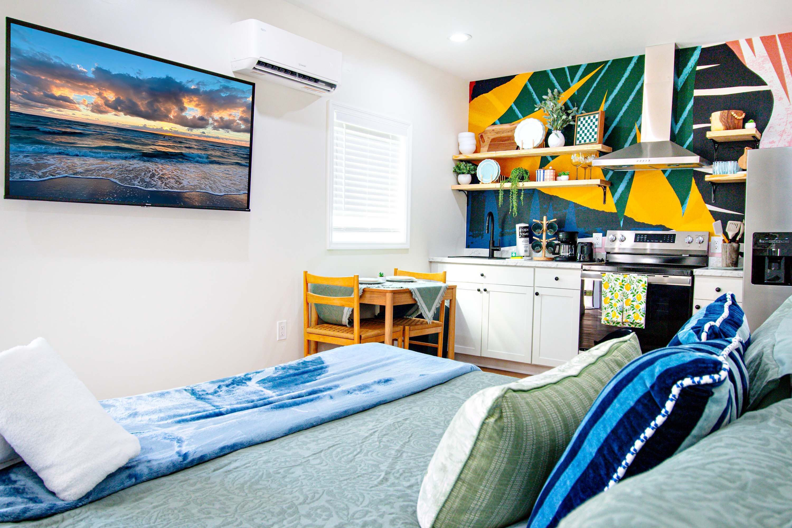 A modern kitchen with a colorful backsplash, a dining area with a table and chairs, a bed with a blue blanket, and a large painting of a sunset.