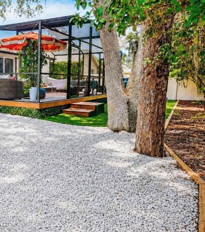A covered outdoor seating area with a gravel ground, a tree, and a patio with steps leading to a building. - Private UCF Apt | King + Priv Yard + Outdoor Tub (Orlando)