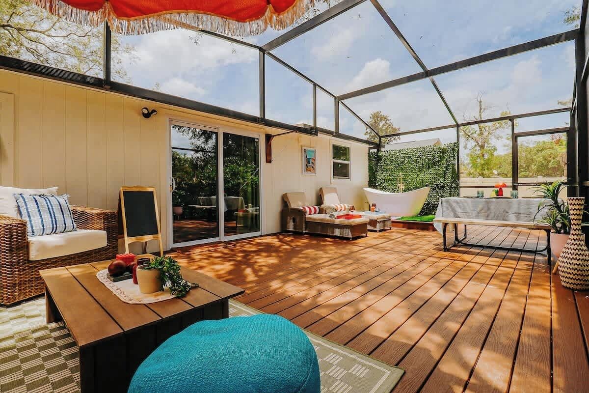 A covered patio with wooden flooring, outdoor furniture, and a glass door leading to a garden.