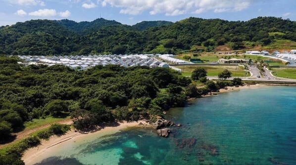 Ett flygbildsbild av ett kustområde med en rad vita byggnader, troligen ett semesterresort eller hotell, omgivet av tät vegetation och ett klart, blått hav.