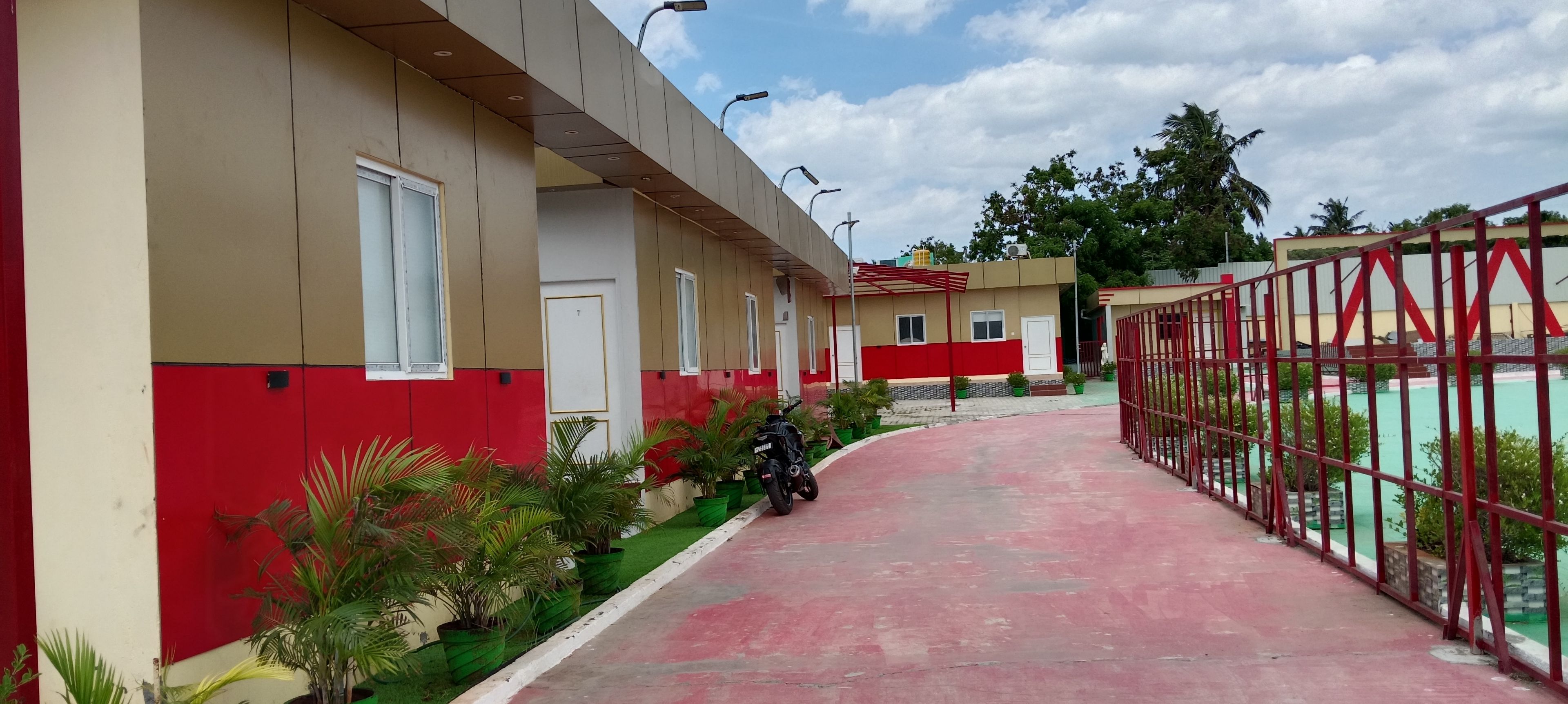 A pathway leading to a building with a red and beige facade, a motorcycle parked beside, and a fenced area with greenery.