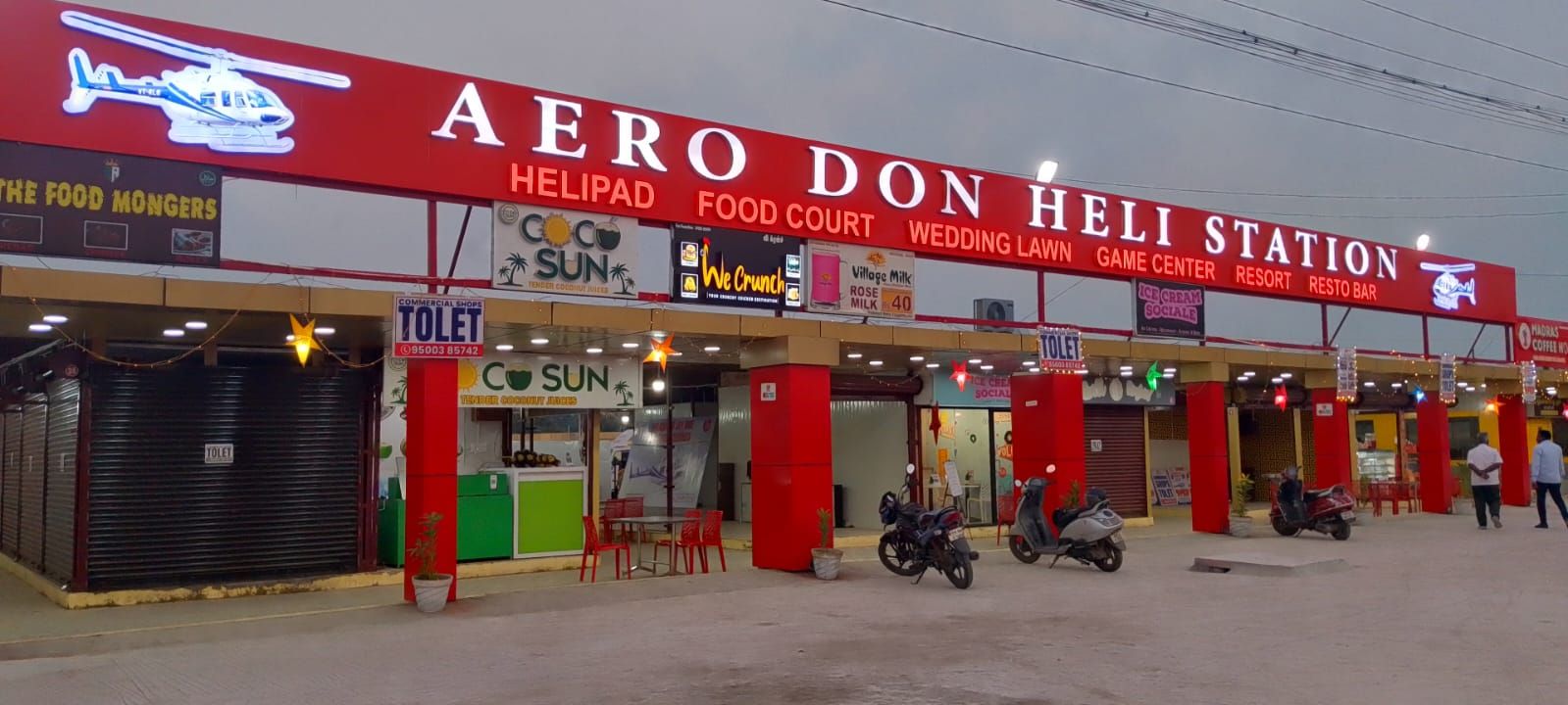 A commercial building with a red and white sign reading 'AERO DON HELI STATION' and various business signs including 'Coco Sun' and 'We Crunch.'